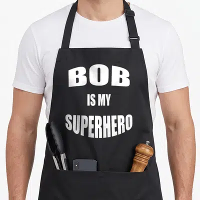 Tablier de cuisine noir avec l'inscription blanche en majuscules BOB IS MY SUPERHERO centrée sur le devant.