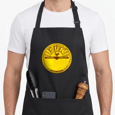 Tablier de cuisine noir avec le logo circulaire jaune et marron de Sun Record Company featuring un coq et des notes de musique.