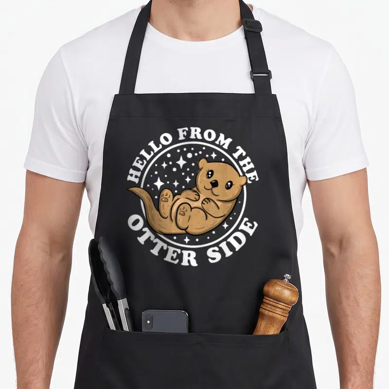 Tablier de cuisine noir avec une illustration de loutre de mer et texte blanc circulaire.