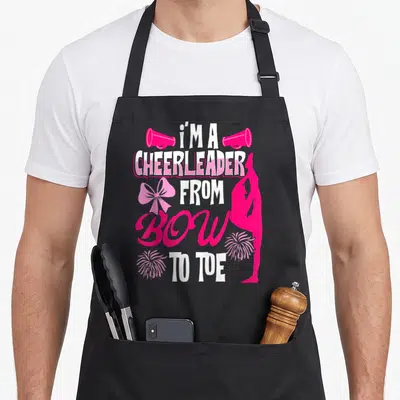 Tablier de cuisine noir avec texte cheerleader bow to toe, pompons roses et silhouette d'acrobate.