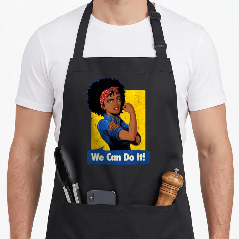 Tablier de cuisine noir avec illustration colorée de Rosie The Riveter et texte We Can Do It en bas.