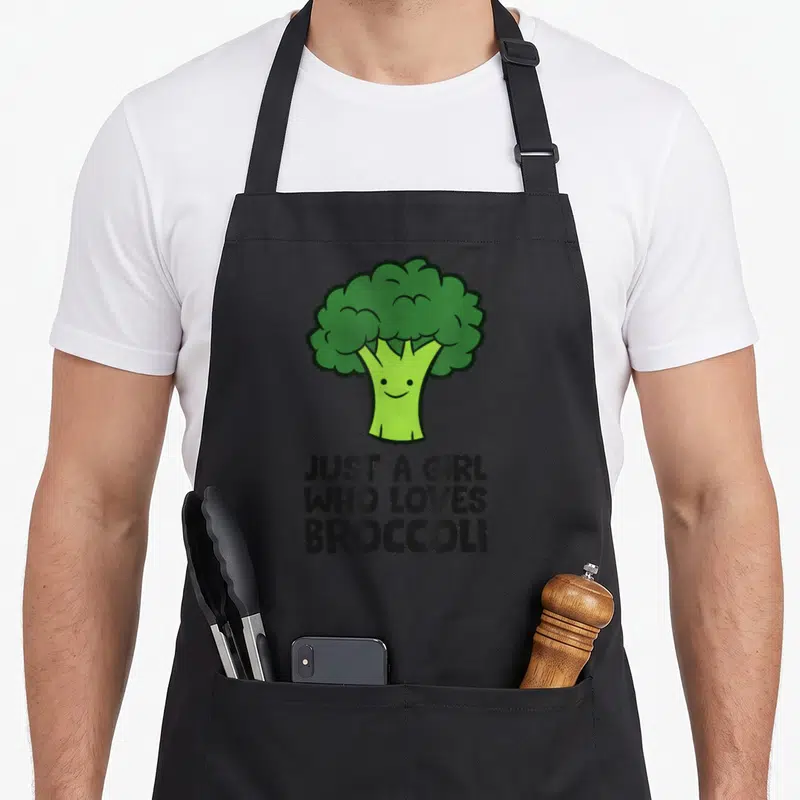 Tablier de cuisine noir avec illustration d'un brocoli vert souriant et texte en anglais sous le dessin.