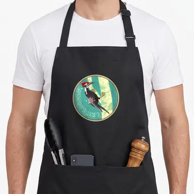 Tablier de cuisine noir avec illustration circulaire d'un woodpecker aux couleurs vives sur fond turquoise.