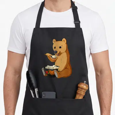 Tablier de cuisine noir avec un ours brun mangeant des pelmeni dans un bol floral traditionnel.