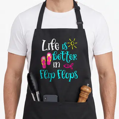 Tablier de cuisine noir avec inscription Life is Better in Flip Flops et illustrations de sandales roses, soleil et poisson.