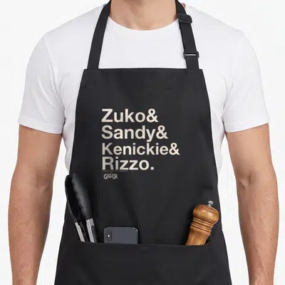 Tablier de cuisine noir avec une pile de noms Zuko Sandy Kenickie Rizzo et le logo Grease en bas.