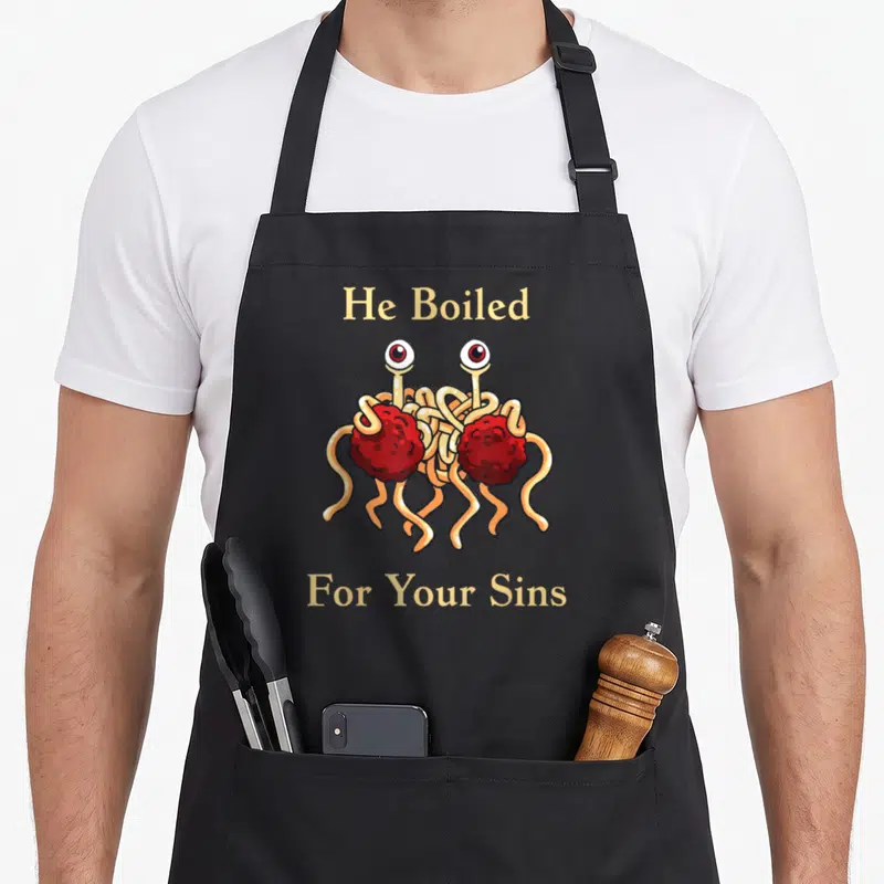 Tablier de cuisine noir avec un monstre de spaghettis aux yeux et la mention He Boiled For Your Sins imprimée en jaune.