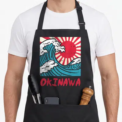 Tablier de cuisine noir avec illustration de vague bleue, soleil rouge rayonnant et texte Okinawa en bas.