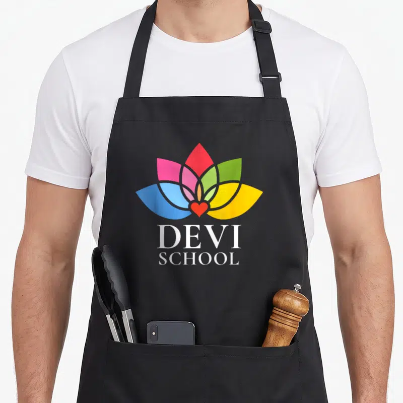 Tablier de cuisine noir avec une fleur aux pétales roses, verts, jaunes et bleus surmontant le texte Devi School en blanc.