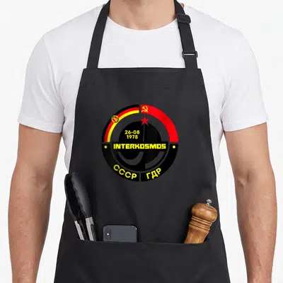 Tablier de cuisine noir avec logo circulaire Interkosmos, drapeaux et inscriptions jaunes sur fond sombre.