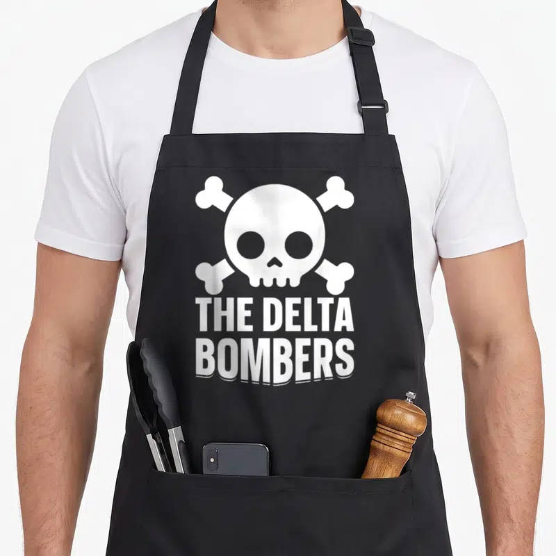 Tablier de cuisine noir avec un crâne blanc, des os croisés et le texte The Delta Bombers imprimé en lettres capitales.