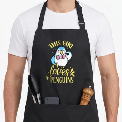 Tablier de cuisine noir avec illustration d'un pingouin bleu portant un nœud papillon rose et le texte This Girl Loves Penguins en jaune.