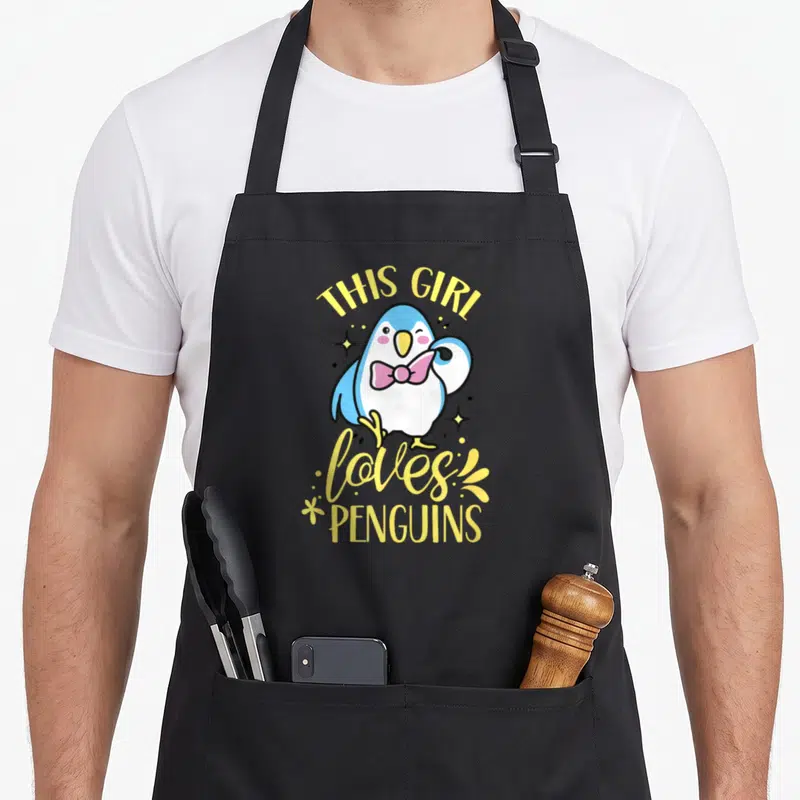 Tablier de cuisine noir avec illustration d'un pingouin bleu portant un nœud papillon rose et le texte This Girl Loves Penguins en jaune.
