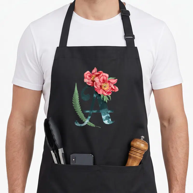 Tablier de cuisine noir avec une grande lettre A décorée de fleurs roses et d'une feuille verte.