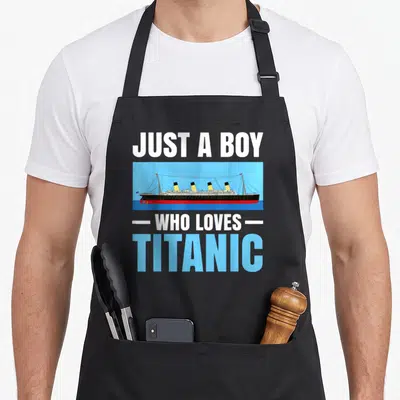 Tablier de cuisine noir avec illustration du paquebot Titanic et le texte Just a boy who loves en typographie blanche et bleue.
