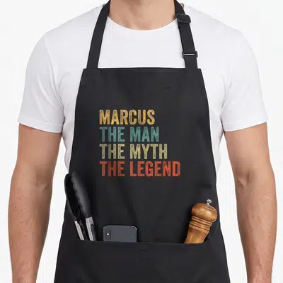 Tablier de cuisine noir avec inscription colorée MARCUS THE MAN THE MYTH THE LEGEND en typographie vintage.