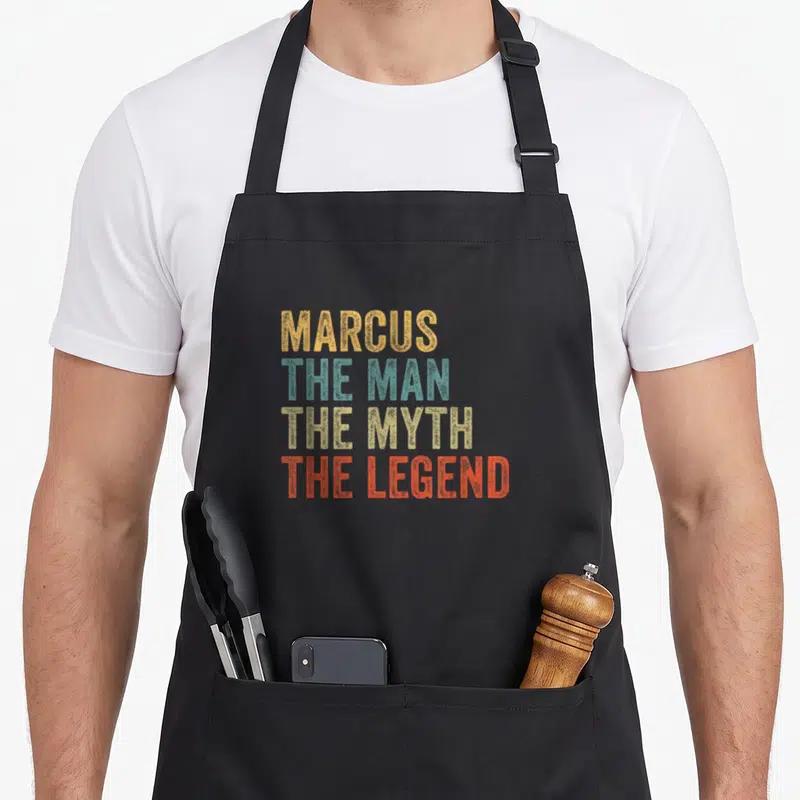 Tablier de cuisine noir avec inscription colorée MARCUS THE MAN THE MYTH THE LEGEND en typographie vintage.