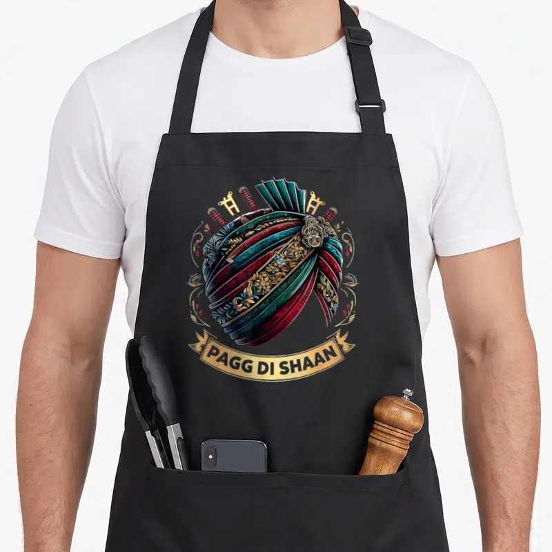 Tablier de cuisine noir avec illustration d'un turban sikh multicolore et l'inscription Pagg di Shaan sur un ruban doré.