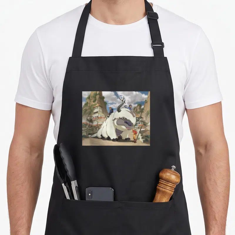 Tablier de cuisine noir avec illustration d'Aang, Appa, Katara, Sokka et Momo devant un village montagnard.