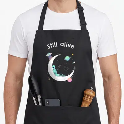 Tablier de cuisine noir avec un alien turquoise assis sur une lune blanche sous le texte still alive.