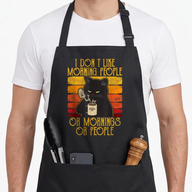 Tablier de cuisine noir avec un chat noir tenant une tasse de café et un texte humoristique en anglais sur fond dégradé orange et rouge.
