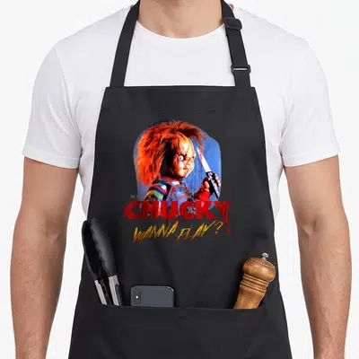 Tablier de cuisine noir avec le portrait effrayant de Chucky tenant un couteau et le texte Wanna Play en rouge.