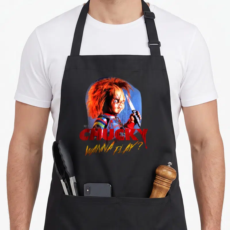 Tablier de cuisine noir avec le portrait effrayant de Chucky tenant un couteau et le texte Wanna Play en rouge.