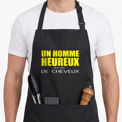 Tablier de cuisine noir avec l'inscription Un homme heureux n'a pas de cheveux en typographie jaune et blanche.