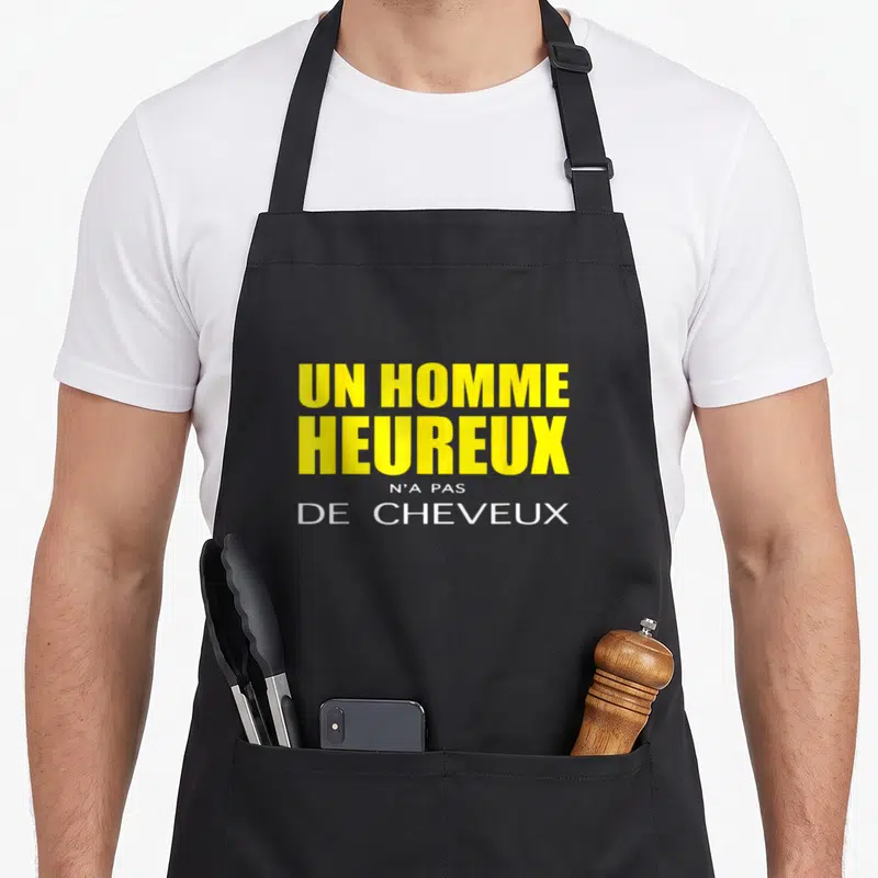 Tablier de cuisine noir avec l'inscription Un homme heureux n'a pas de cheveux en typographie jaune et blanche.
