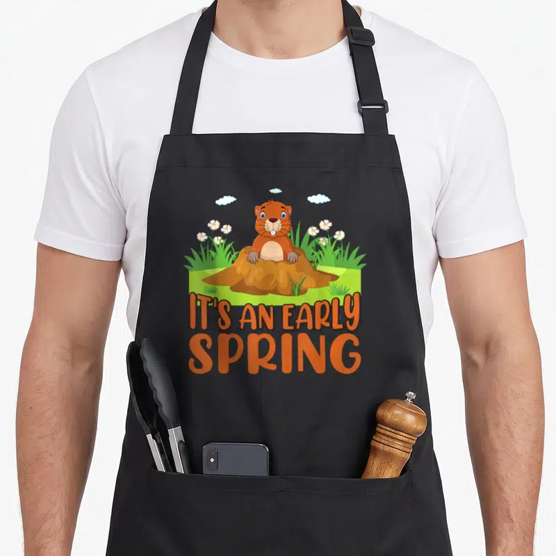 Tablier de cuisine noir avec un taupier cartoon sortant d'un terrier entouré de fleurs et le texte It's an early spring en orange.