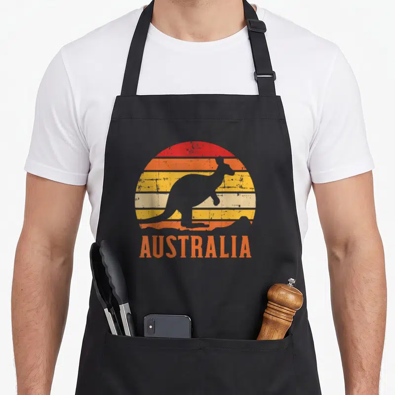 Tablier de cuisine noir avec silhouette de kangourou et texte AUSTRALIA sur fond de soleil rayé orange et jaune.