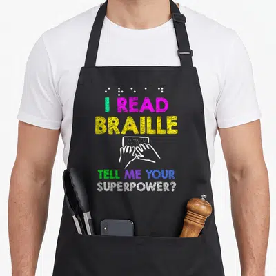 Tablier de cuisine noir avec l'inscription I READ BRAILLE TELL ME YOUR SUPERPOWER en lettres colorées et des mains lisant le pointé.
