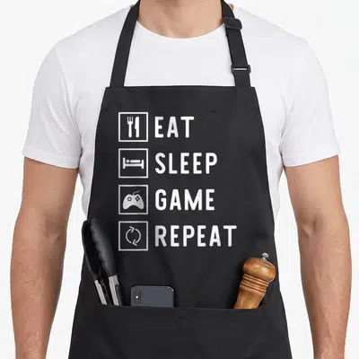 Tablier de cuisine noir avec les mots Eat Sleep Game Repeat et quatre icônes blanches alignées verticalement.