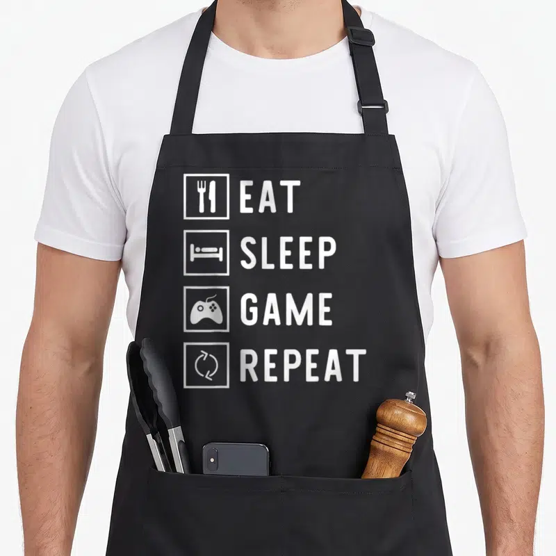 Tablier de cuisine noir avec les mots Eat Sleep Game Repeat et quatre icônes blanches alignées verticalement.