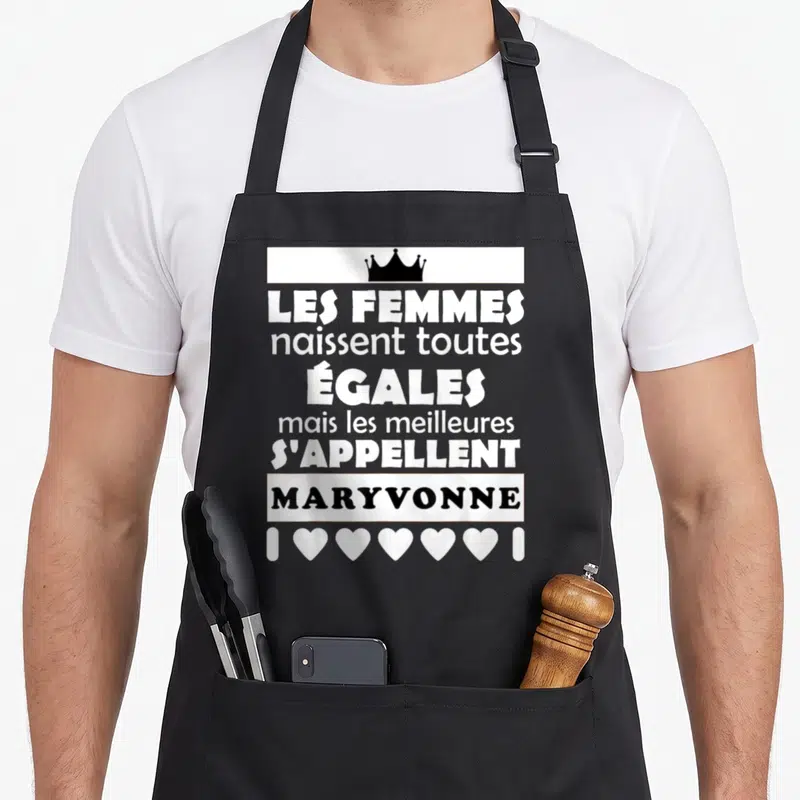 Tablier de cuisine noir avec citation blanche Les femmes naissent toutes égales et le prénom Maryvonne en noir sous une couronne.