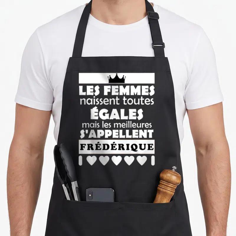 Tablier de cuisine noir avec citation Les femmes naissent toutes égales Frédérique, couronne et cœurs blancs.
