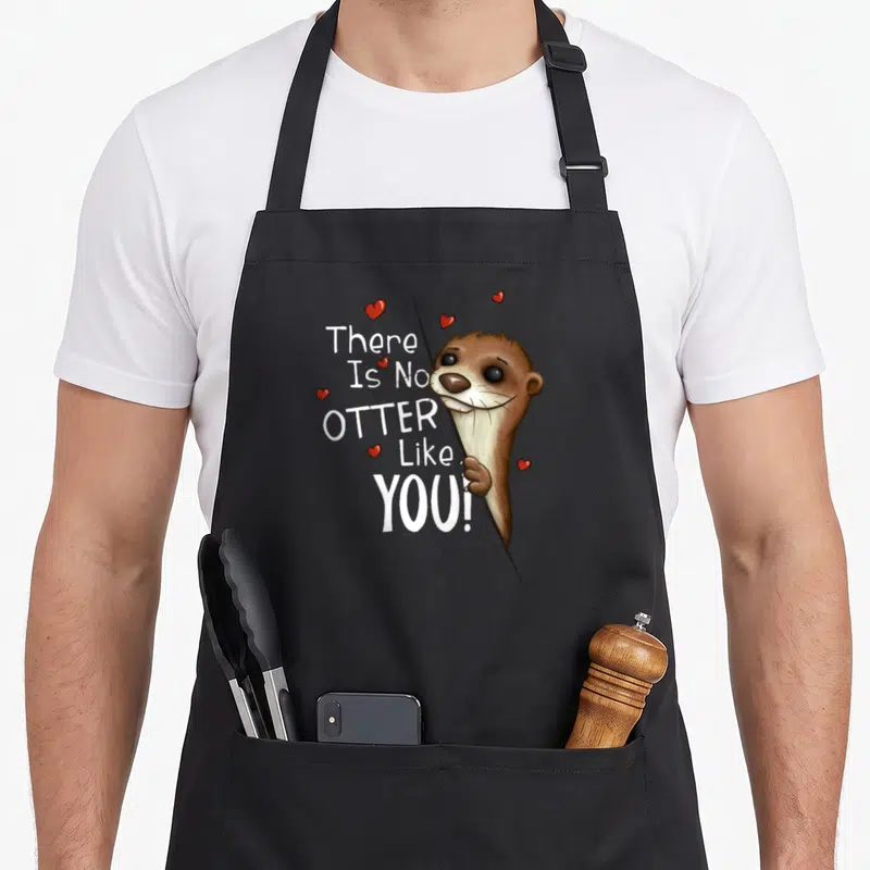 Tablier de cuisine noir avec une loutre marron et le texte There Is No Otter Like You entouré de petits cœurs rouges.