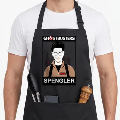 Tablier de cuisine noir avec portrait stylisé de Spengler, logo Ghostbusters et nom en lettres blanches sur fond gris uni.