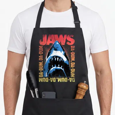 Tablier de cuisine noir avec illustration de requin Jaws et texte Da Dum en couleurs vives sur fond sombre.