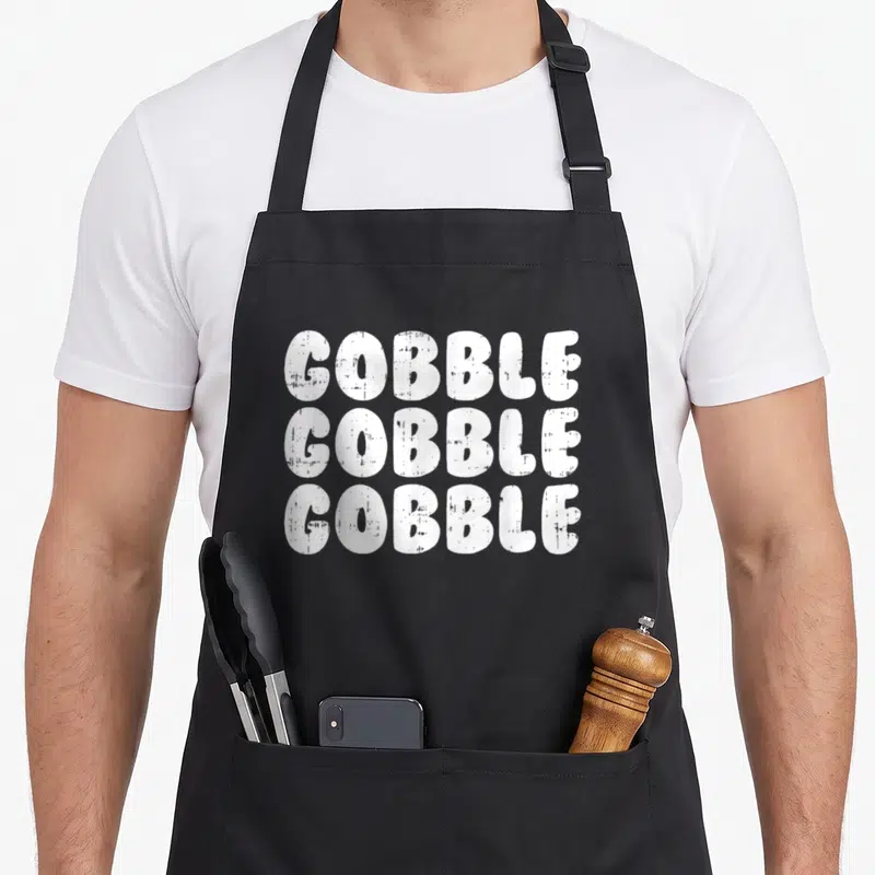 Tablier de cuisine noir avec l'inscription Gobble Gobble Thanksgiving Turkey Day en lettres blanches épaisses et texturées.