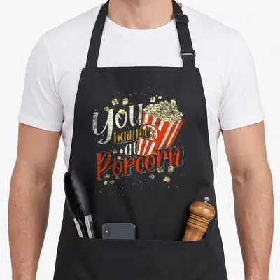 Tablier de cuisine noir avec l'inscription Tu M'As Eu Au Popcorn et une barquette de popcorn vintage.