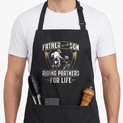 Tablier de cuisine noir avec illustration père et fils motards et texte en anglais.