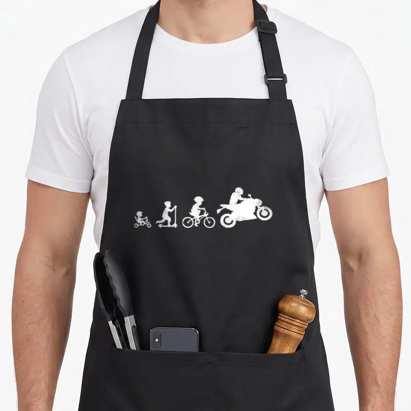 Tablier de cuisine noir avec une illustration blanche montrant la progression d'un enfant vers un pilote de moto.