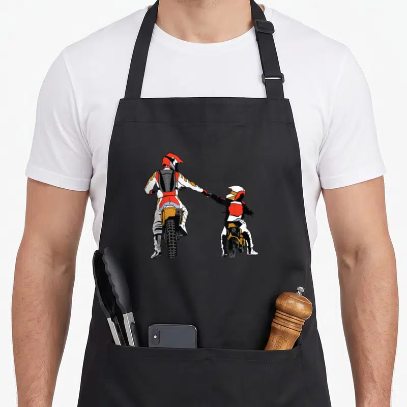 Tablier de cuisine noir avec illustration d'un pilote de moto et un enfant en tenue de cross se tenant la main de dos.