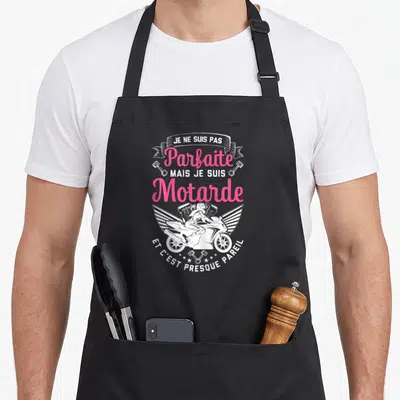 Tablier de cuisine noir avec une illustration rose et blanche d'une motarde ailée et des pistons.