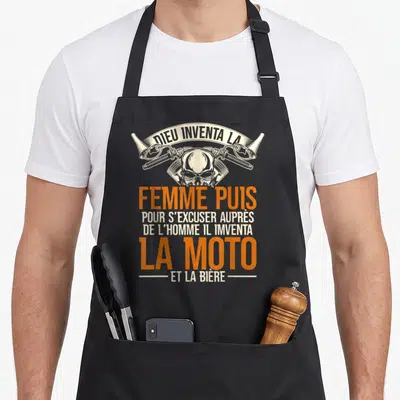 Tablier de cuisine noir avec un crâne, des pistons croisés et le texte Dieu inventa la femme puis la moto et la bière en orange et blanc.