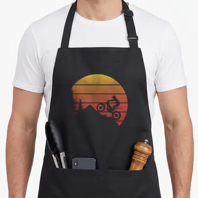 Tablier de cuisine noir avec illustration circulaire d'un pilote de trial sur fond de soleil orange strié et silhouettes d'arbres.