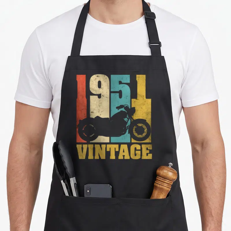 Tablier de cuisine noir avec motif vintage 1951 et silhouettes de motos colorées sur fond texturé.