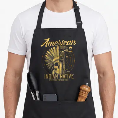 Tablier de cuisine noir avec un skull coiffé de plumes et une roue de moto imprimés en doré au centre.