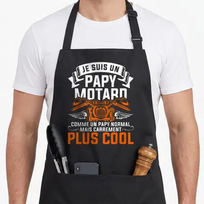Tablier de cuisine noir avec inscription Je suis un papy motard et moteur orange stylisé aux ailes blanches.